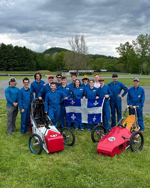 L'équipe est sur un terrain de course automobile et sourient à la caméra en tennant un drapeau du Québec devant leurs bolides de courses