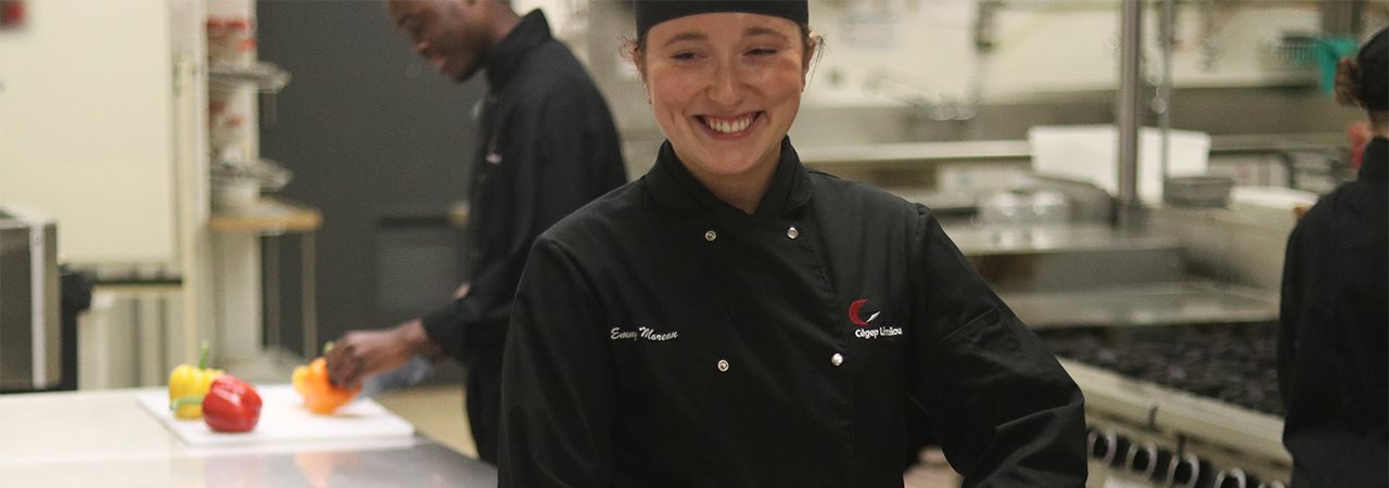 Étudiante souriante en cuisine, et étudiant en arrière-plan qui coupe des poivrons