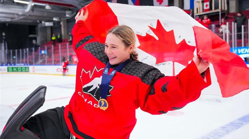 Marilou tiens le drapeau du canada en patinant sur le glace dans son uniforme de hockey canada.